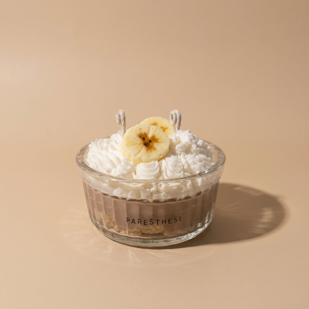 A dessert in a clear ribbed glass bowl with the brand name PARESTHESI visible on the front, topped with whipped cream and a banana slice, set on a warm beige background.