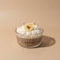A dessert in a clear ribbed glass bowl with the brand name PARESTHESI visible on the front, topped with whipped cream and a banana slice, set on a warm beige background.