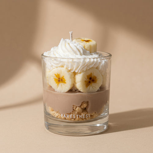 A clear glass jar candle with a white whipped-cream-like top and banana slices inside, labeled PARESTHESI, on a light beige surface.