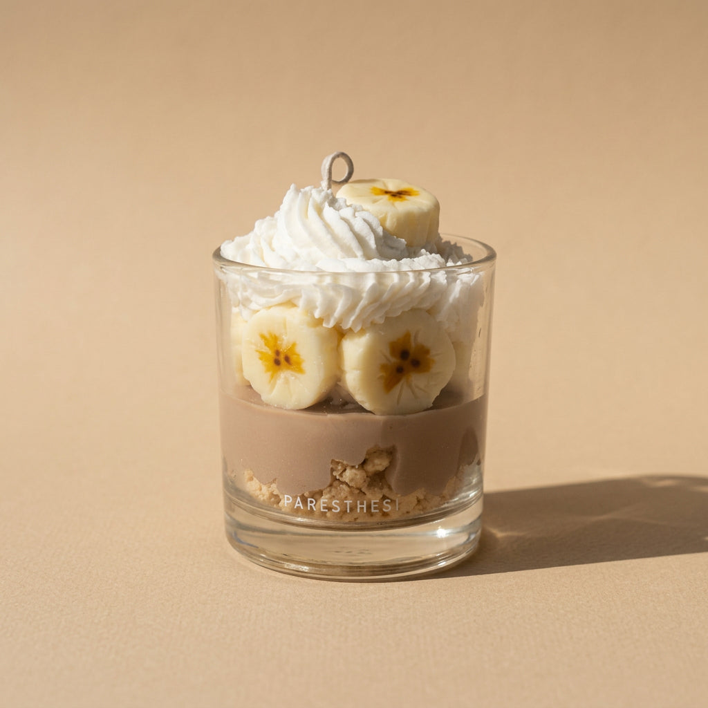 A clear glass jar candle with a lid, filled with creamy white whipped wax and sliced banana pieces, on a light beige background. The glass side is printed with the brand name 'PARESTHESI'.