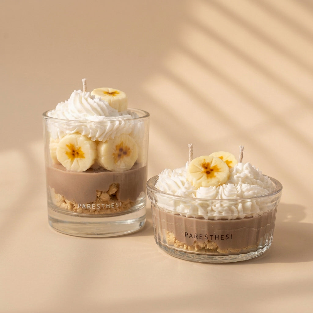Two clear glass jar candles with white whipped cream and banana slices, set against a light, neutral background. One candle has a white label reading 'PARESTHESI,' and the other has a black label with the same brand.