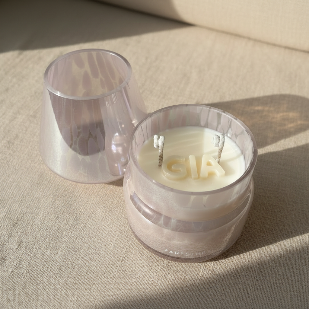 Candle with 'Gia' spelled out in white wax, inside a pink pearl lamp jar on a beige fabric background.