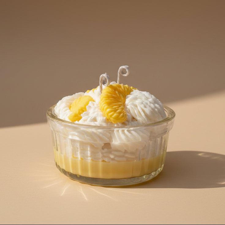 A candle that looks like a dessert in a glass bowl with whipped cream and lemon slices on a beige background
