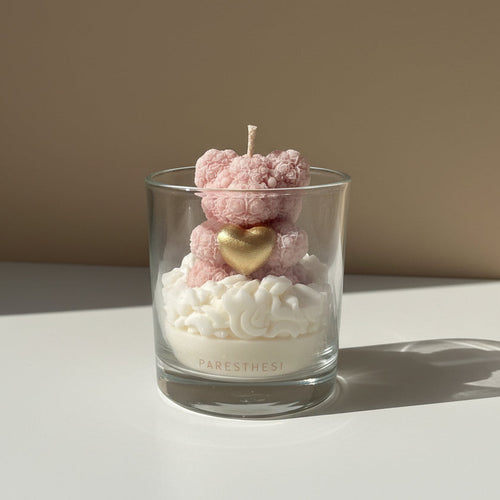 Candle shaped like a pink teddy bear with a gold heart inside a glass container on a white surface.