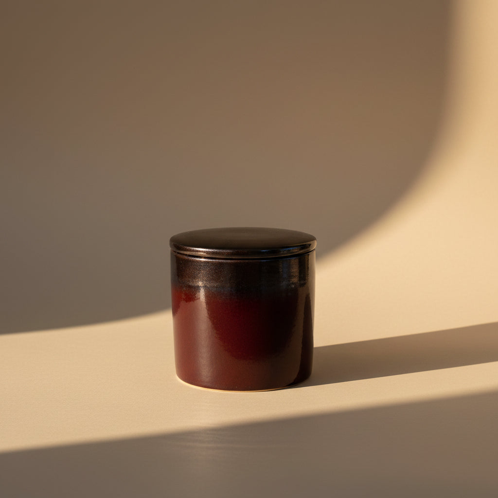 A small cylindrical ceramic container with a matching lid, featuring a glossy dark reddish-brown glaze and a contrasting dark gray glazed top, shown against a plain off-white background.