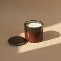 A ceramic candle jar with a white wax surface and two cotton wicks, shown next to its matching metallic lid on a warm beige background.