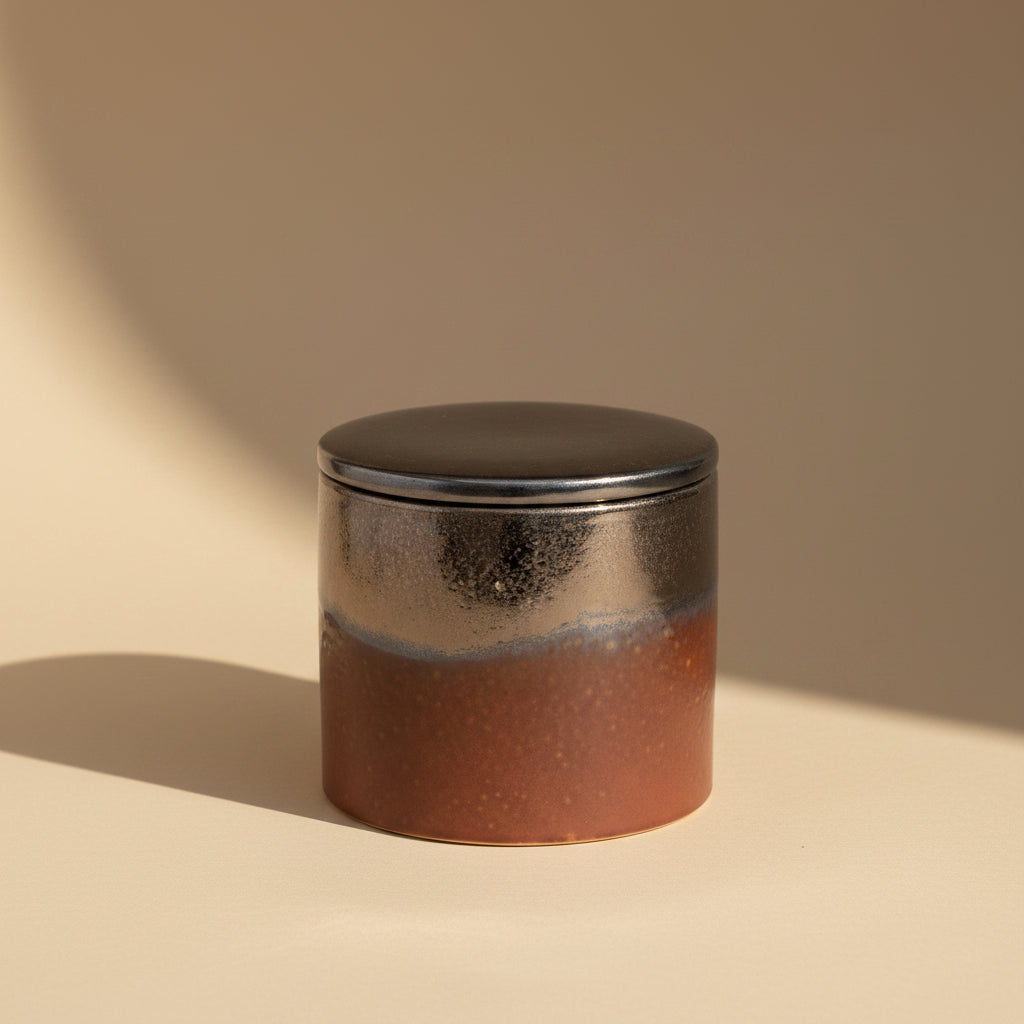 A small cylindrical ceramic jar with a matching lid, featuring a glossy metallic silver upper section and a textured dark red-brown lower section, set against a plain white background.