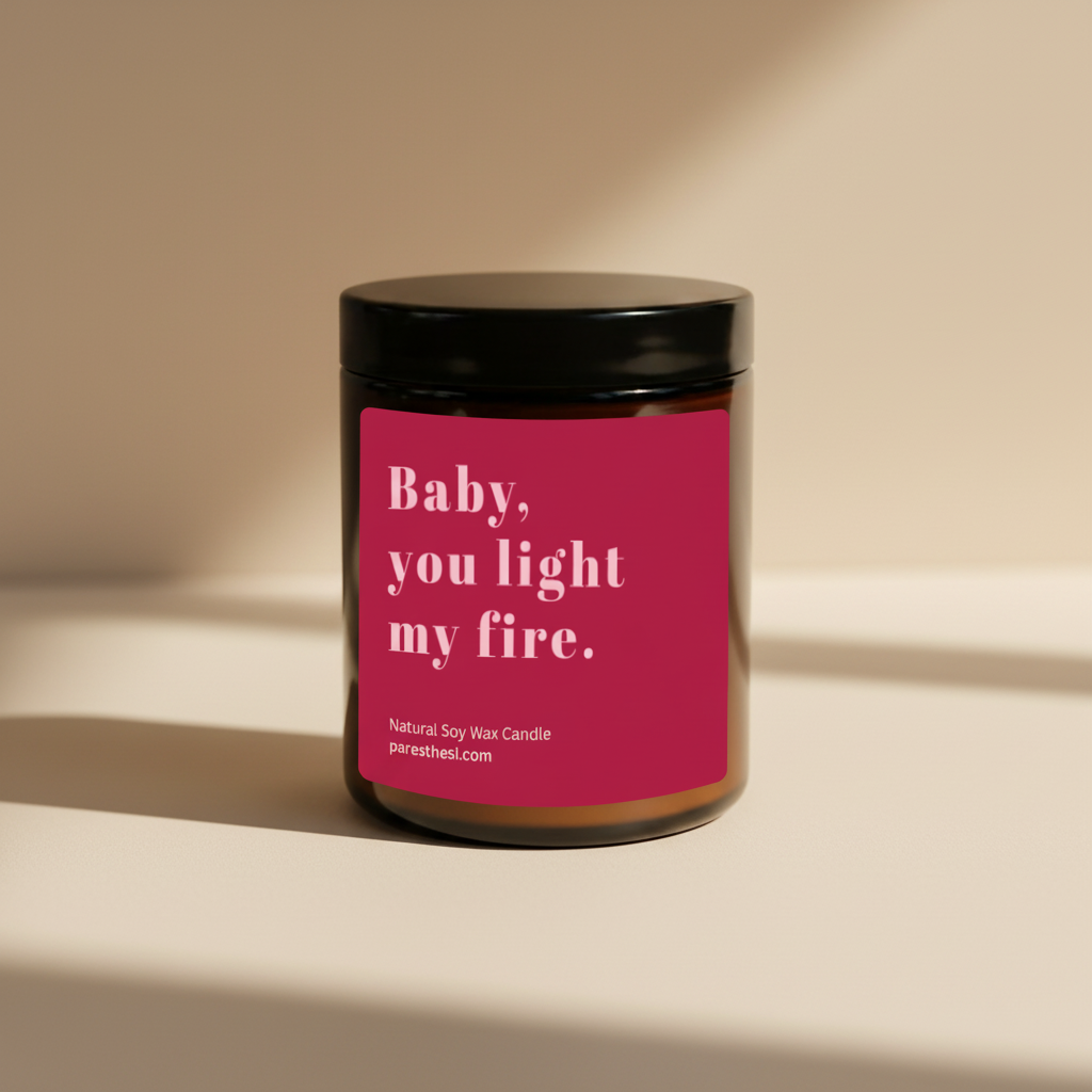 A glass amber jar candle with a black lid and a bright red label that reads 'Baby, you light my fire.' in light pink text, shown on a plain beige background.