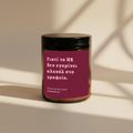 A single amber jar candle with a black lid and a deep magenta label featuring light pink Greek text, set against a beige background.