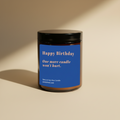A dark amber glass jar candle with a black lid and a blue label that reads 'Happy Birthday. One more candle won't hurt.' in light orange color on a beige background.