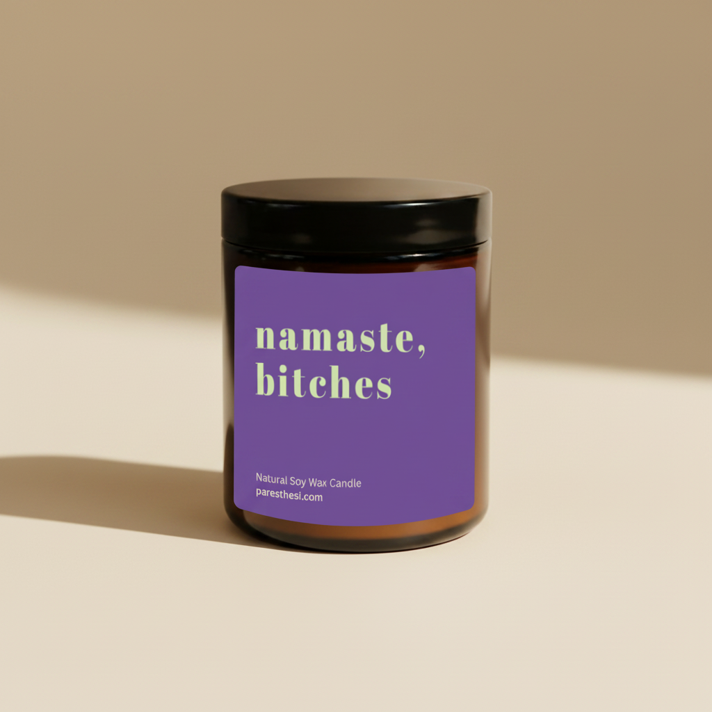 A soy wax candle in an amber glass jar with a black lid and a purple label that reads 'namaste, bitches' in pale green text, shown on a plain beige background.