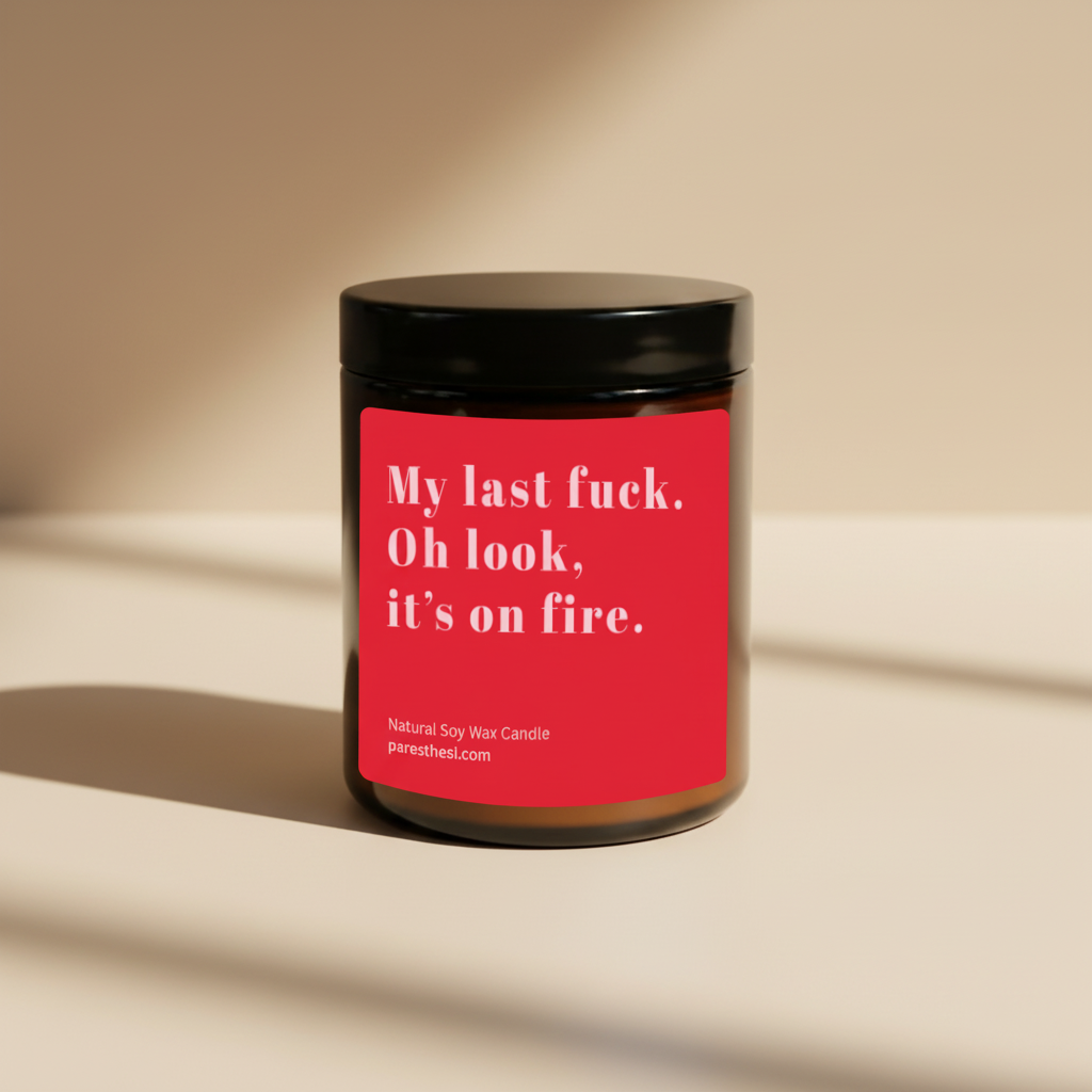 A jar candle with a black lid and a bright red label featuring white text that reads, 'My last fuck. Oh look, it's on fire.