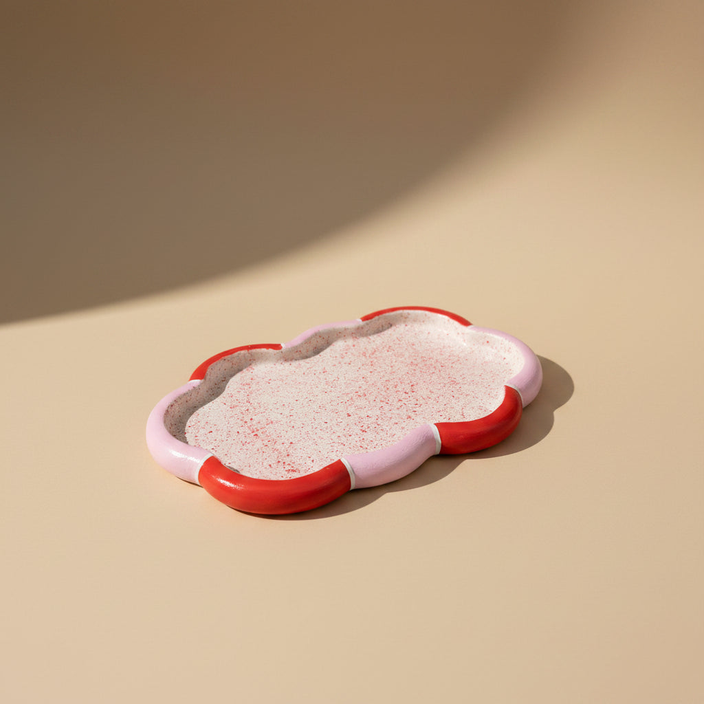 A small cloud-shaped porcelain tray with a speckled white interior and a glossy red and pale pink exterior, photographed on a warm beige surface with soft shadows.