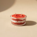 A small clear glass candle jar with two wicks, filled with layered red and white wax resembling a red velvet cake, on a warm beige background. The brand name 'PARESTHESI' is printed on the front of the glass.