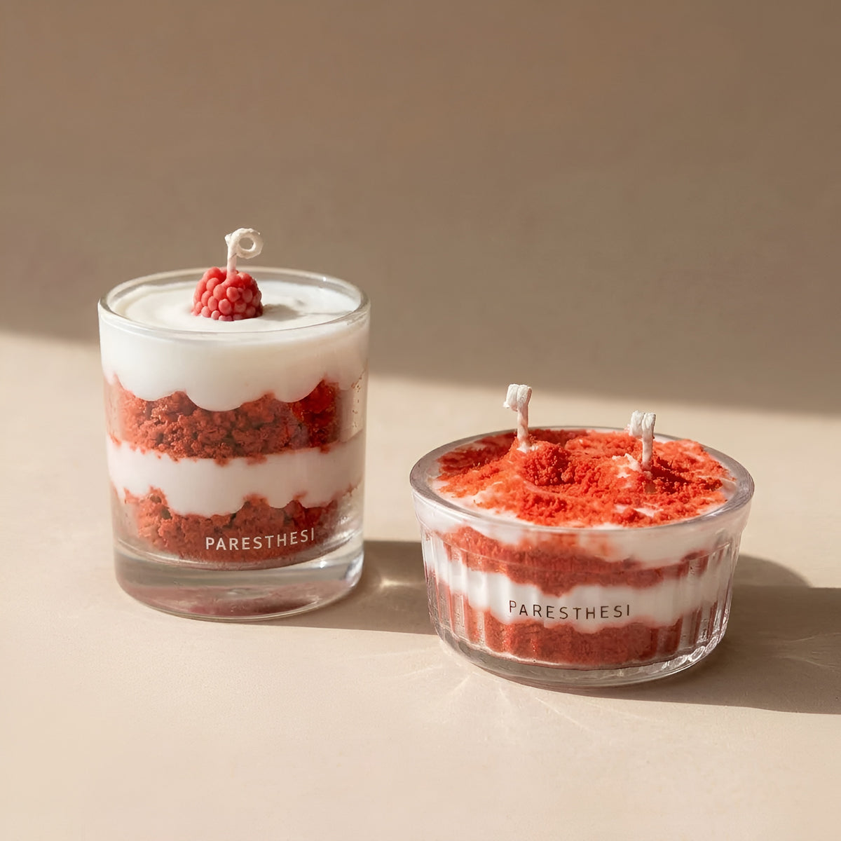 Two layered candles designed to look like red velvet cake layers in clear glass containers, with white wax on top and a small raspberry garnish on one candle. The brand name 'PARESTHESI' is printed on the front of the glassware.