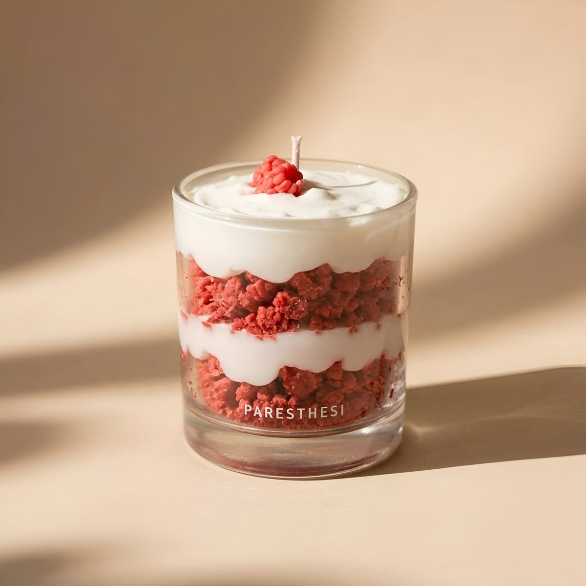 A layered candle in a clear glass jar with red and white layers resembling red velvet cake, topped with white wax and a small red berry, on a beige background with soft shadows. The brand name 'PARESTHESI' is printed on the glass.