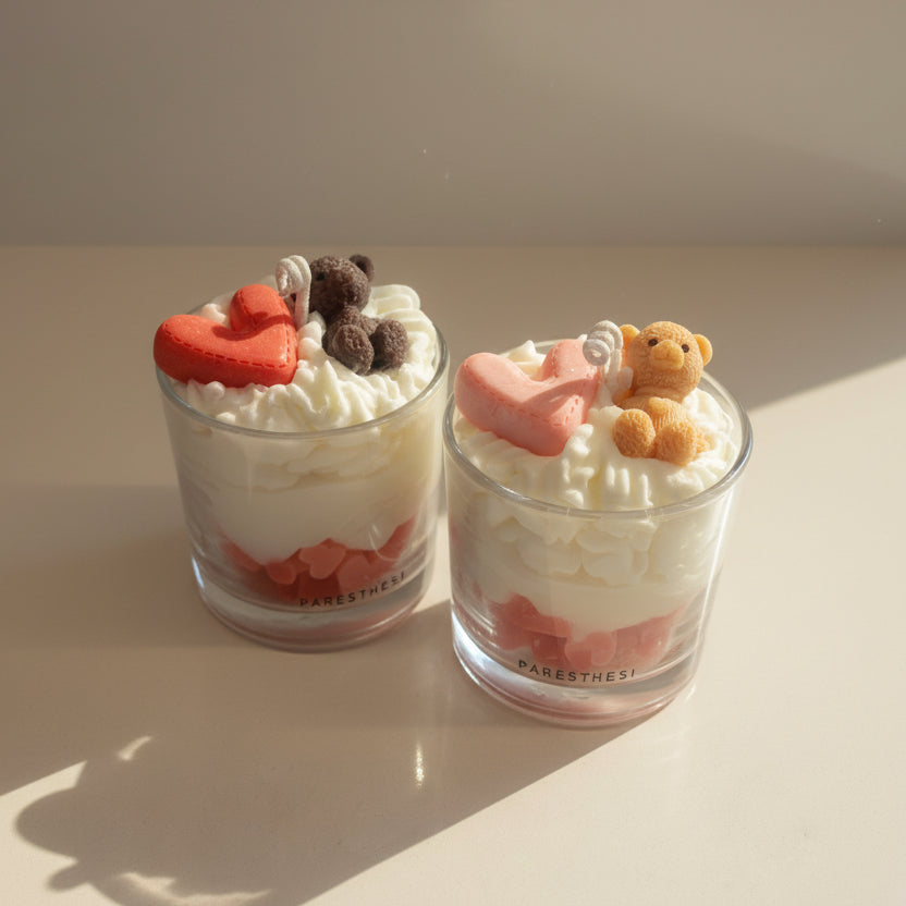 Two candles glasses with whipped cream, strawberries, and small teddy bear and heart decorations on a light background.
