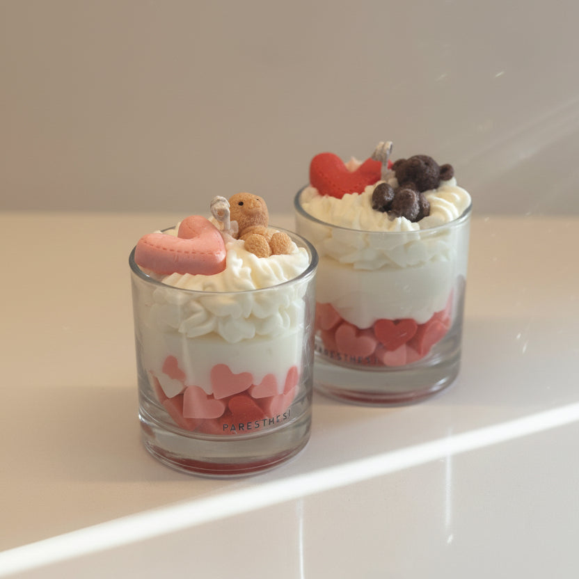 Two glass candles with whipped cream wax, heart-shaped, and small teddy bears on a white surface.