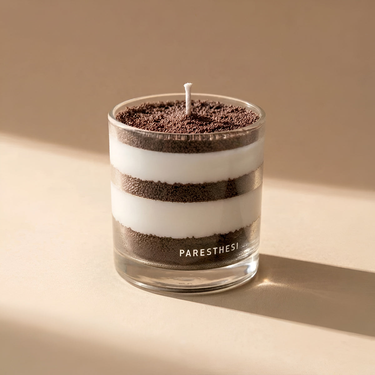 A candle in a clear glass jar with alternating dark brown and white horizontal layers, topped with dark brown crumb-like material and a single cotton wick. The brand name 'PARESTHESI' is printed in white on the lower half of the glass.
