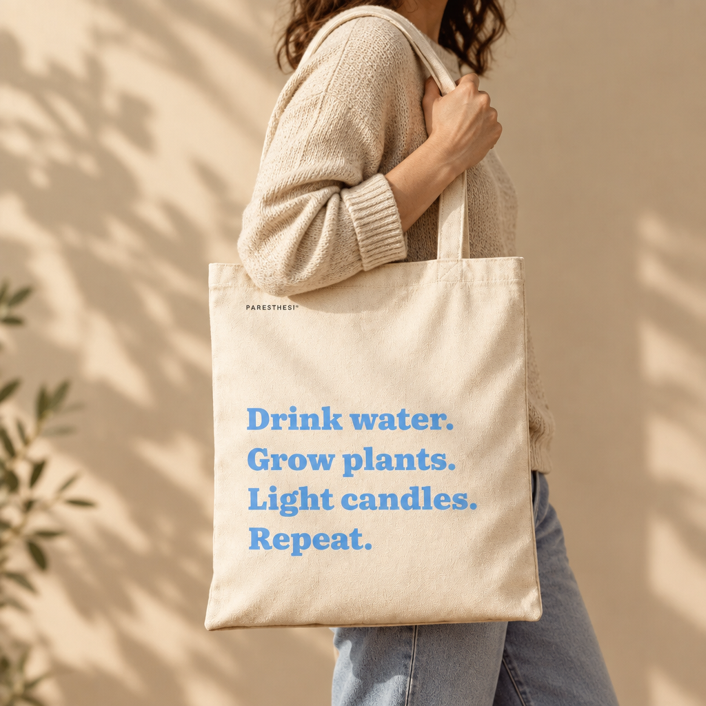 A person wearing a beige knit sweater and light blue jeans carries a large beige canvas tote bag with blue text that reads, 'Drink water. Grow plants. Light candles. Repeat'.
