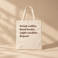A cream canvas tote bag with brown text reading 'Drink coffee. Read books. Light Candles. Repeat'.
