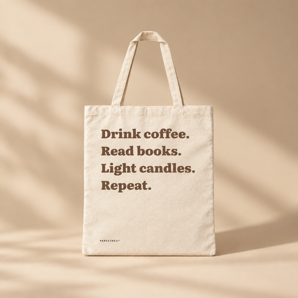 A cream canvas tote bag with brown text reading 'Drink coffee. Read books. Light Candles. Repeat'.