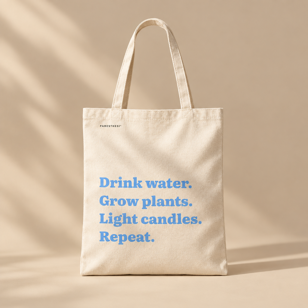 A natural canvas tote bag with light blue printed text that reads 'Drink water. Grow plants. Light Candles. Repeat'.