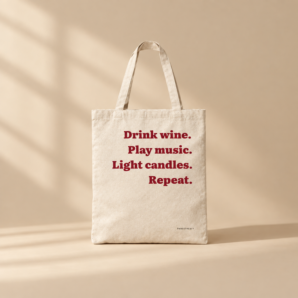A natural canvas tote bag with red printed text that reads 'Drink wine. Play music. Light Candles. Repeat'.