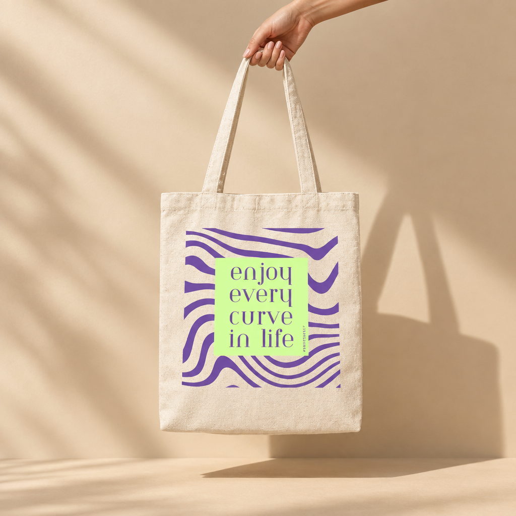 A hand holds a cream canvas tote bag with purple wavy stripes and a central lime green square that reads 'enjoy every curve in life' in purple text, set against a warm beige wall with soft shadows.