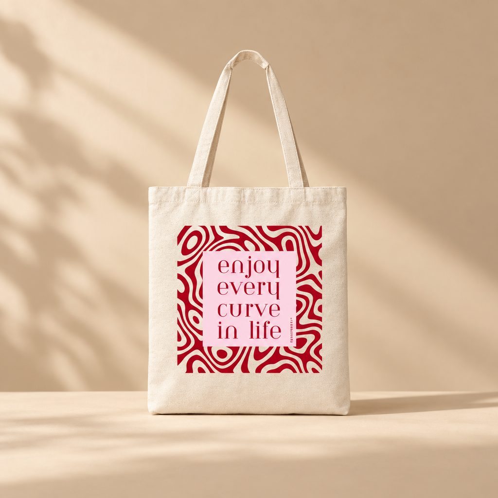 An off-white canvas tote bag with two handles, featuring a square graphic print on the front. The print has a light pink background with the words 'enjoy every curve in life' in dark red text, surrounded by a red wavy pattern.