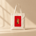 An off-white canvas tote bag with two handles, featuring a prominent red rectangular graphic on the front with Mona Lisa's face peeking out. The bag is photographed on a warm beige surface with soft diagonal shadows cast across it.