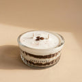 A small, clear glass jar candle with a white wax fill and two wicks, decorated with dark coffee beans and cocoa powder on top. The side of the jar is printed with the brand name 'PARESTHESI'.