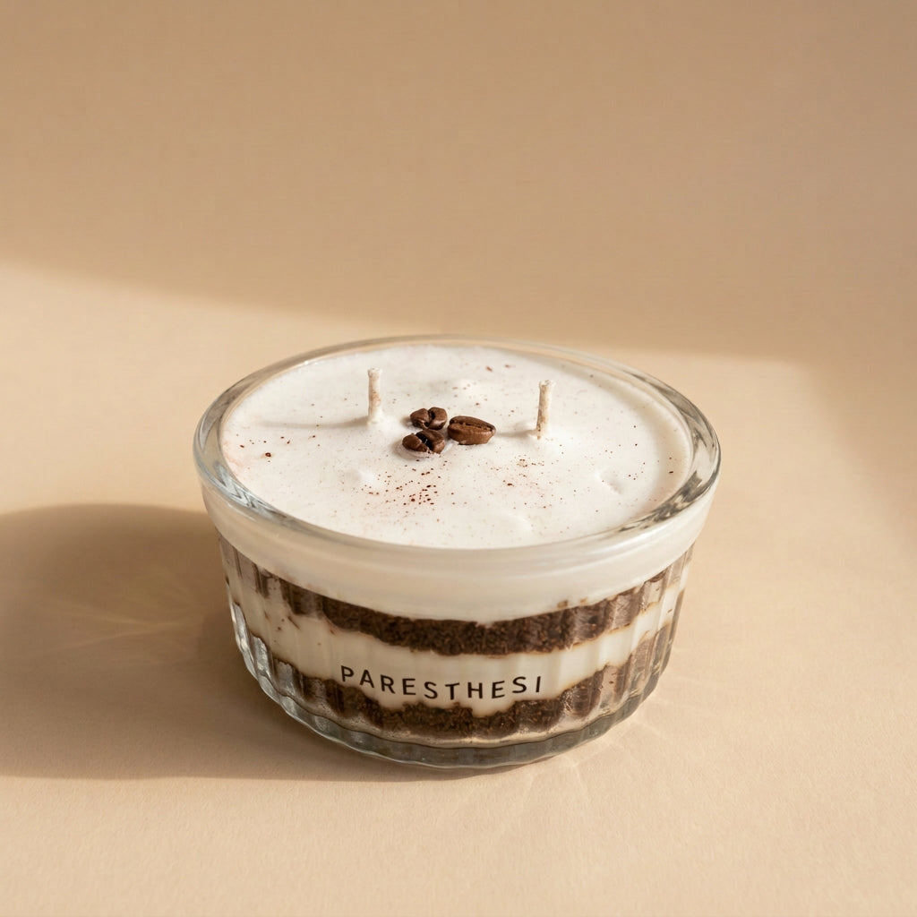 A small, clear glass jar candle with a white wax fill and two wicks, decorated with dark coffee beans and cocoa powder on top. The side of the jar is printed with the brand name 'PARESTHESI'.