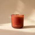 A cylindrical red frosted glass candle with two cotton wicks, standing vertically on its side against a plain beige wall. The brand name is 'PARESTHESI'.