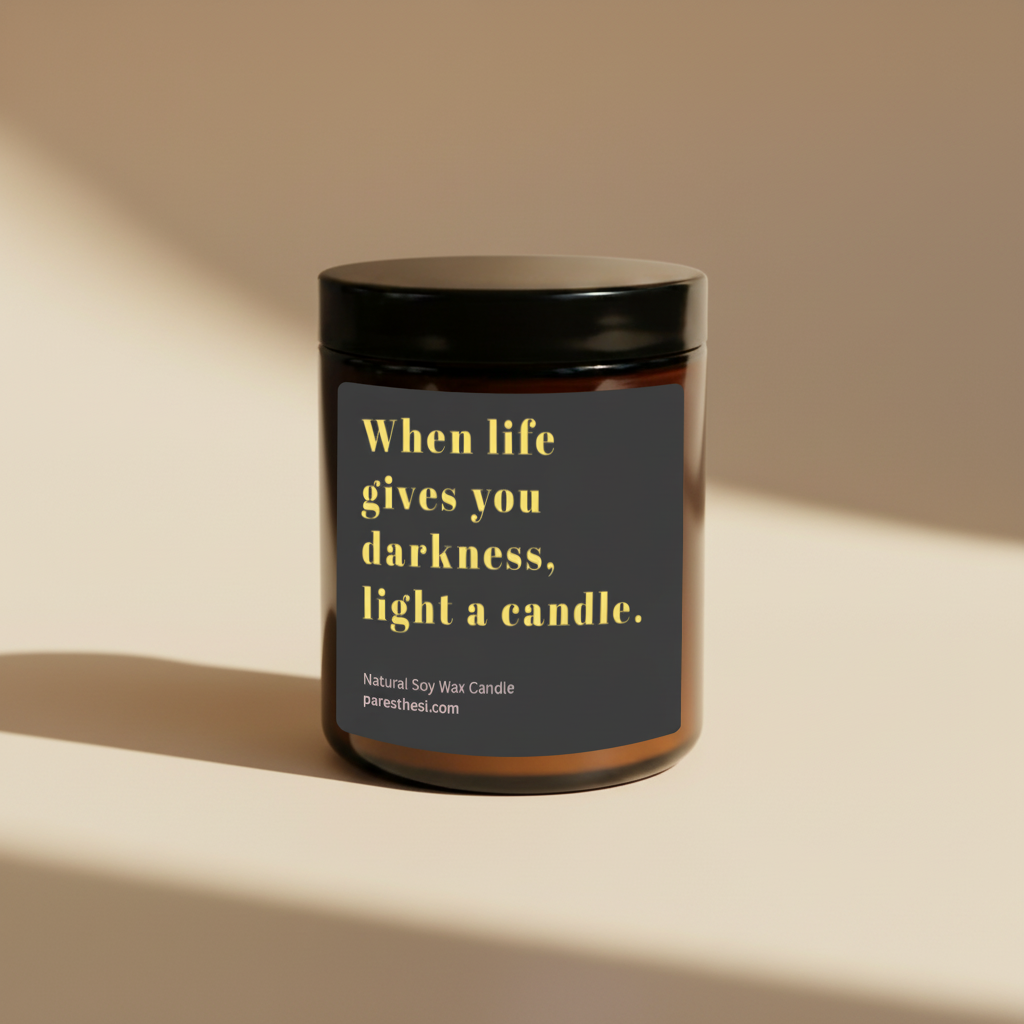 An amber glass jar candle with a black lid and a dark gray label featuring yellow text that reads, 'When life gives you darkness, light a candle.' Smaller text at the bottom reads 'Natural Soy Wax Candle' and 'paresthesi.com'.